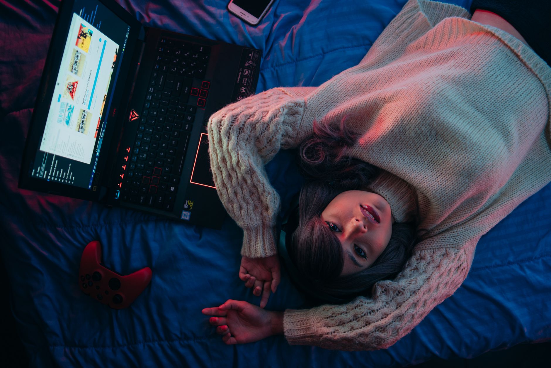 Femme allongée sur un lit, vue de dessus, près d’un ordinateur portable de jeu ouvert et d’une manette rouge, sous une lumière rouge et bleue.