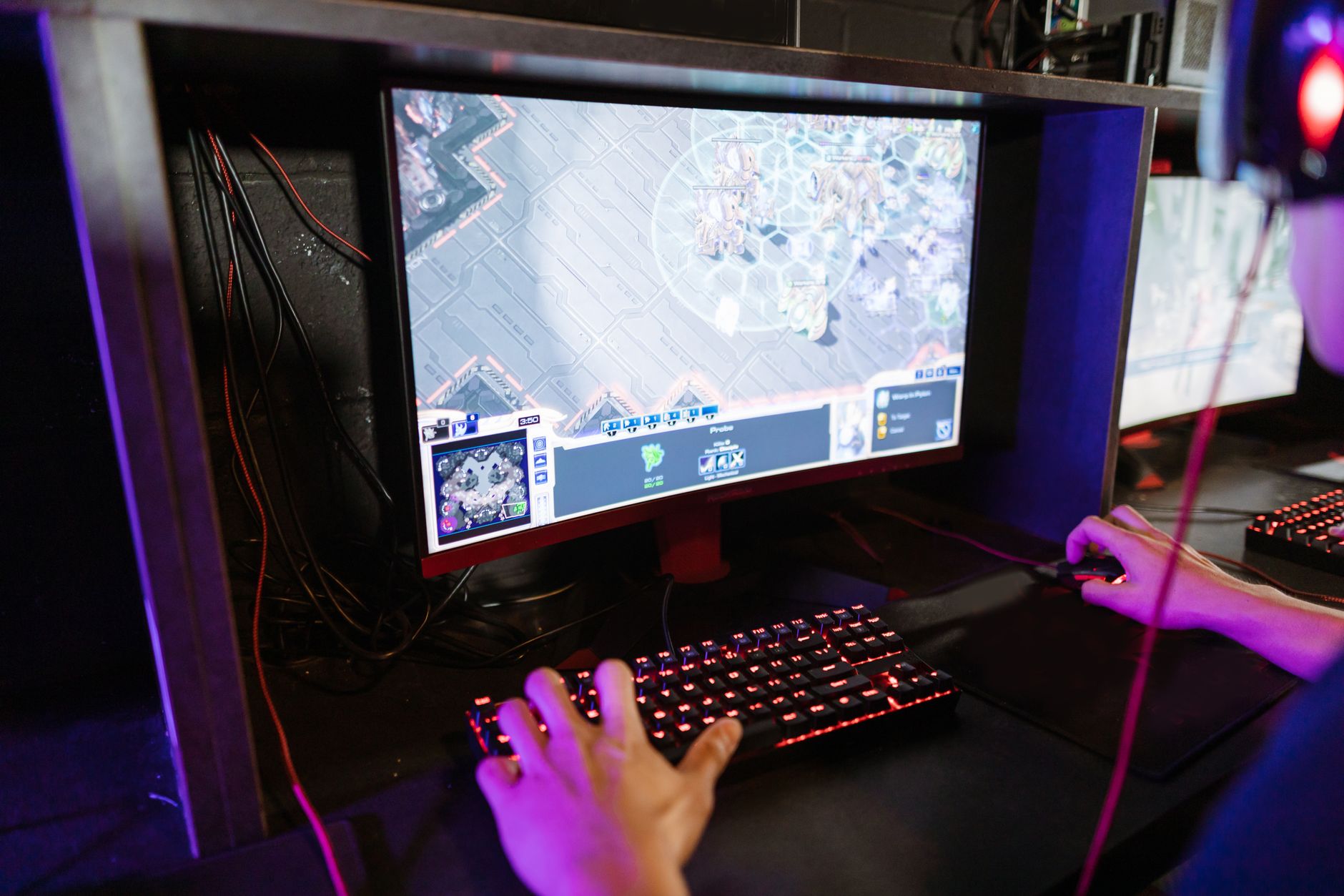 Joueur casqué devant un écran incurvé affichant un jeu de stratégie, une main sur un clavier rétroéclairé rouge, l’autre sur la souris, dans une pièce sombre.