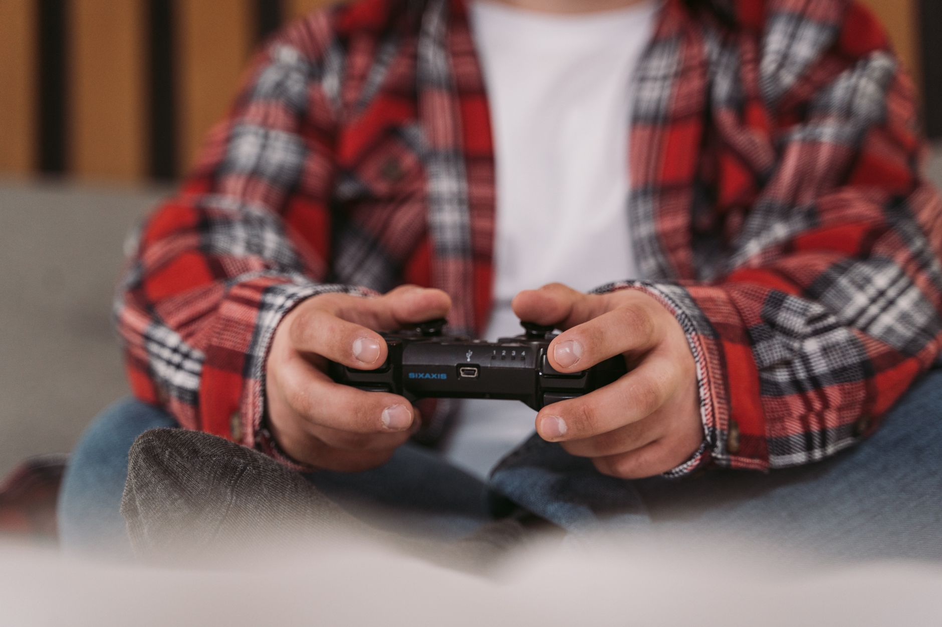 Mains tenant une manette de jeu noire, personne en chemise à carreaux rouge assise sur un canapé dans un intérieur.