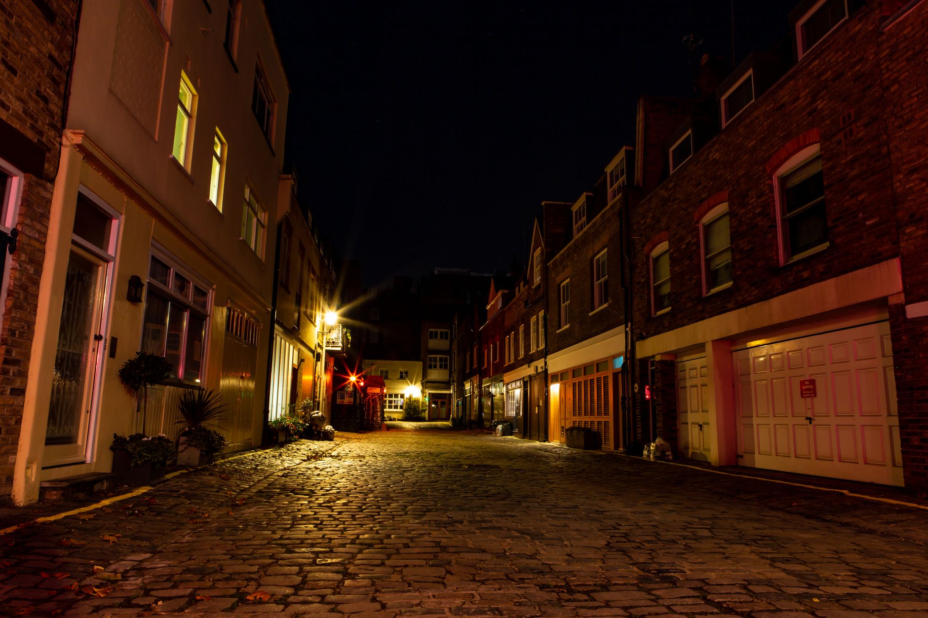 Ruelle pavée déserte la nuit, bordée de maisons en brique et de garages, éclairée par des lampadaires et des fenêtres.