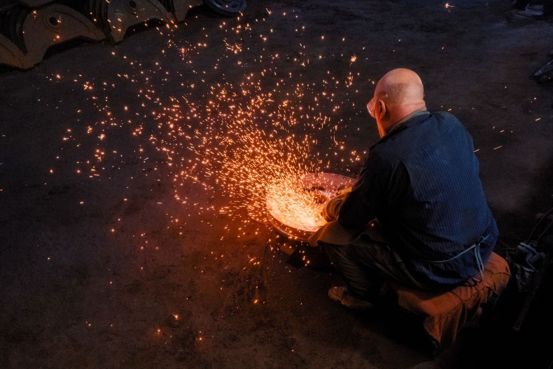 Un homme accroupi dans un atelier sombre travaille une pièce métallique circulaire, entouré d’une gerbe d’étincelles orange.