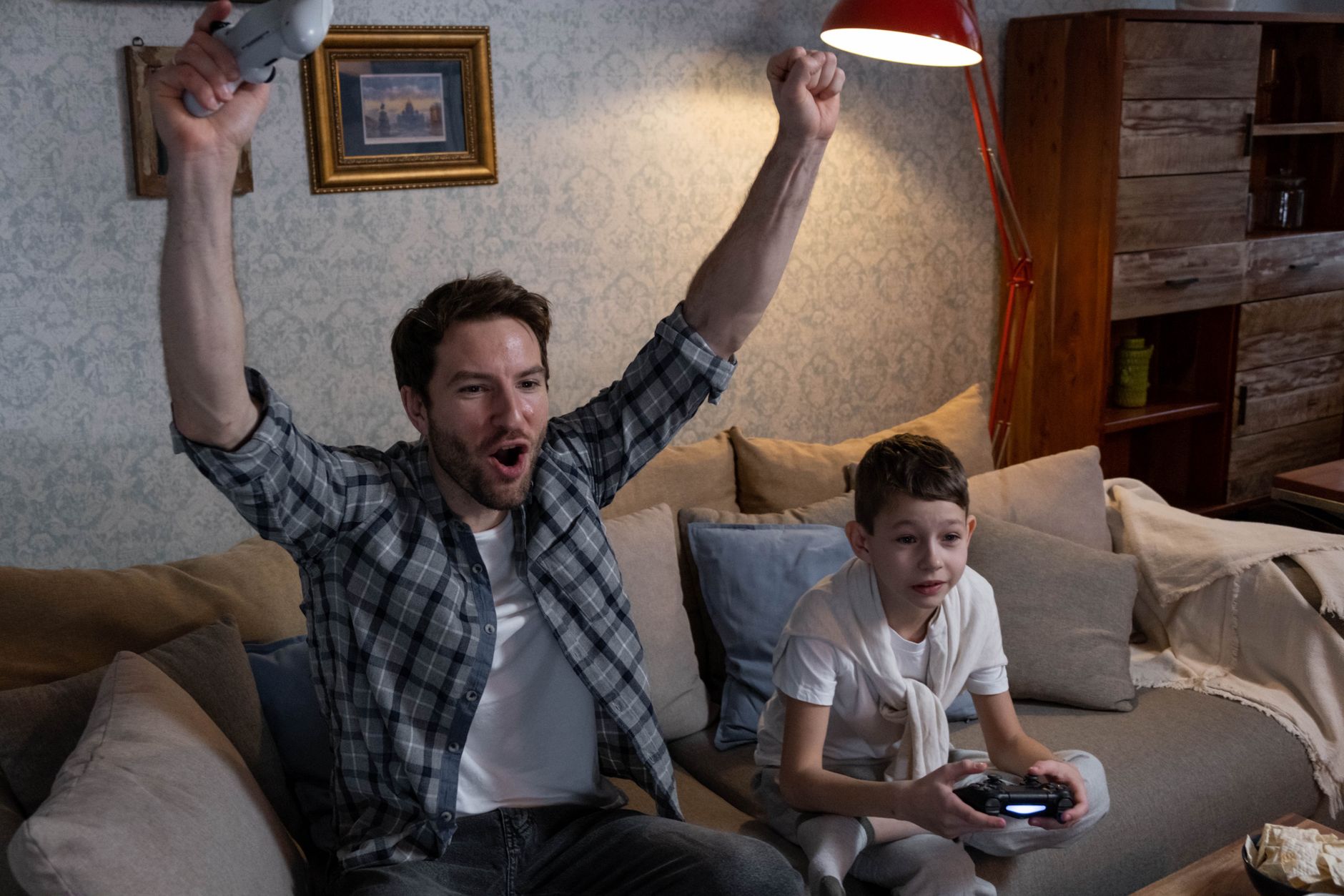 Un homme levant les bras avec une manette et un enfant jouant à un jeu vidéo sur un canapé dans un salon.